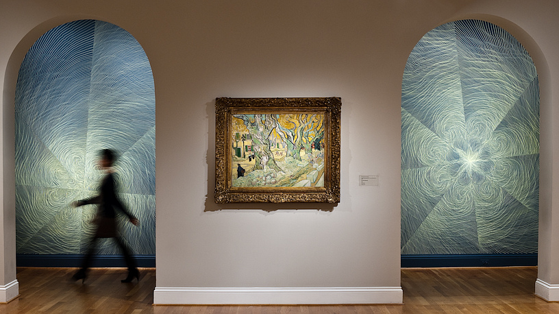 Photo of a figure walking in front of walls with drawings by linn meyers, with a painting by Van Gogh on display