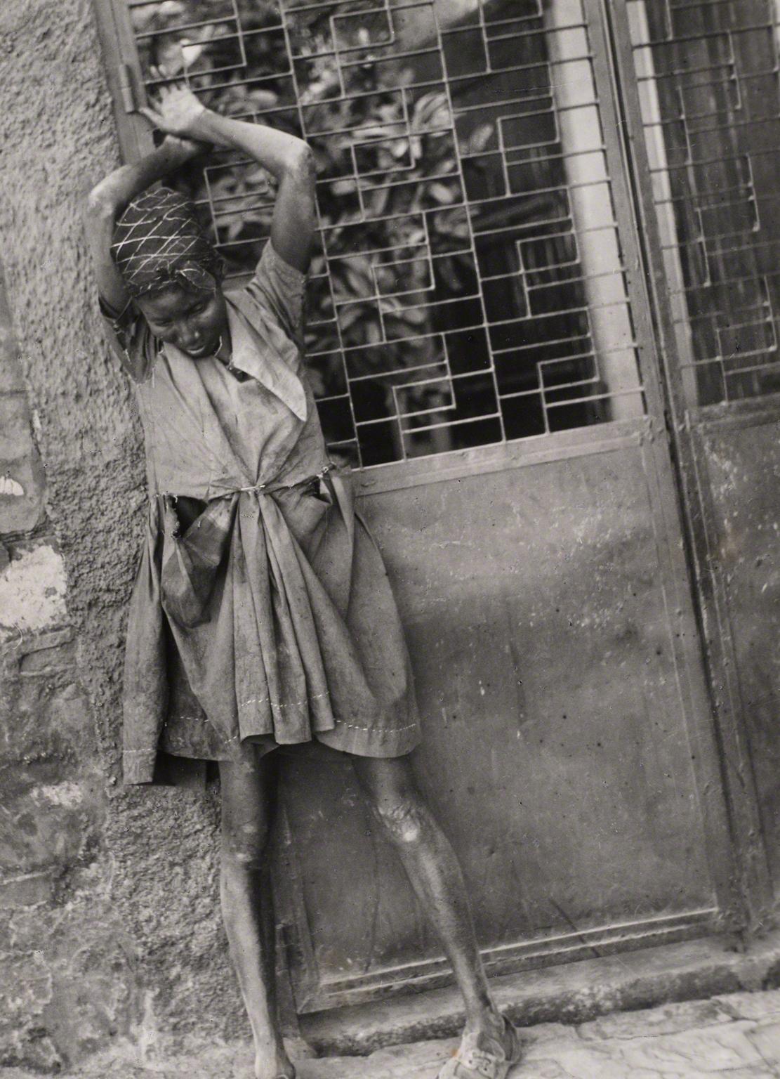 Leon Levinstein, Untitled (Haiti, girl with ragged clothes and skinny legs standing against door with arms up), not dated.